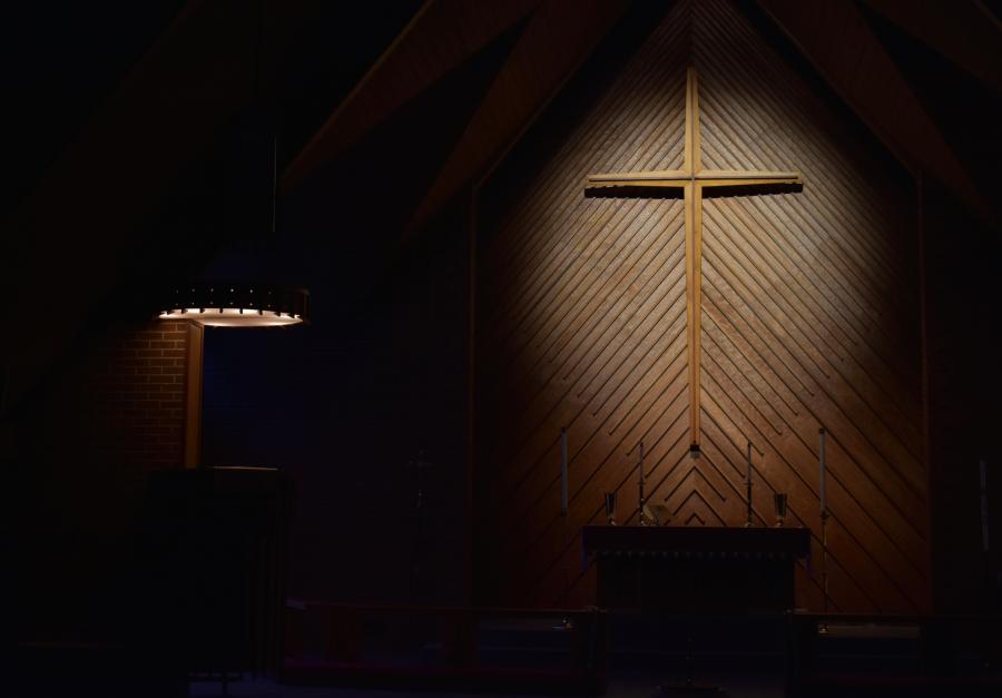 Dark church stage with a spotlight on a cross with a wooden artistic patterned wall behind it
