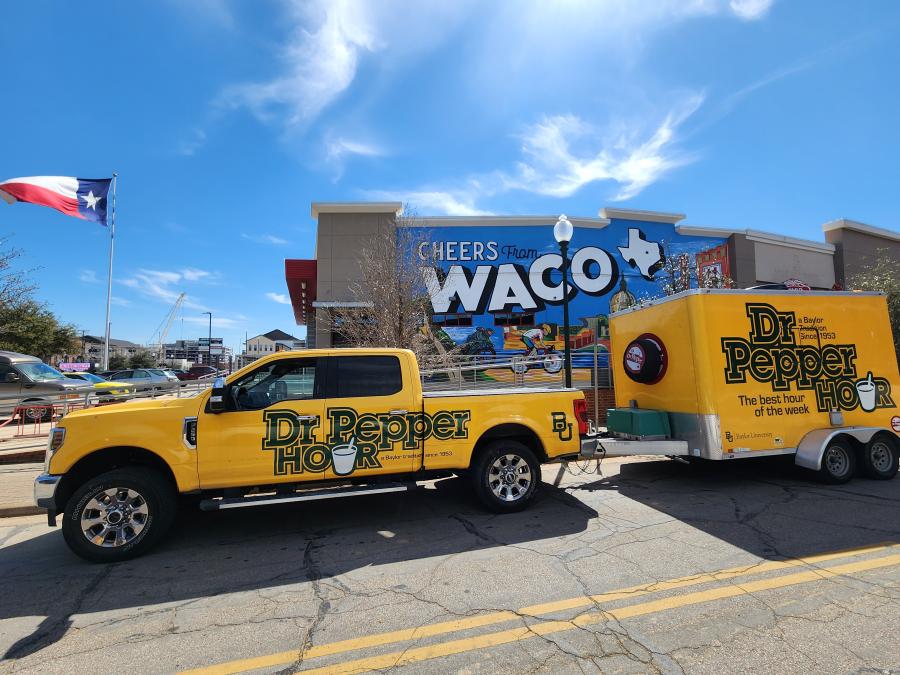 A gold Dr Pepper Hour Tour-Baylor co-branded truck with a trailer parked in front of a building with a Waco mural on the side. There is a large Texas flag in the background and a clear blue sky overhead.