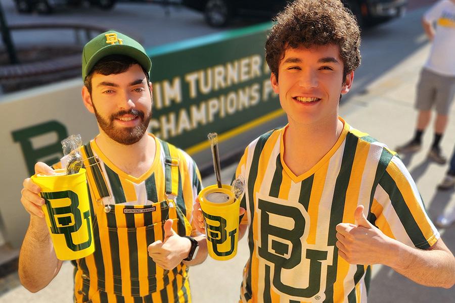 Two Baylor students wearing Bear Pit jerseys striped green, gold and white; holding yellow cups with a green Baylor logo on the front